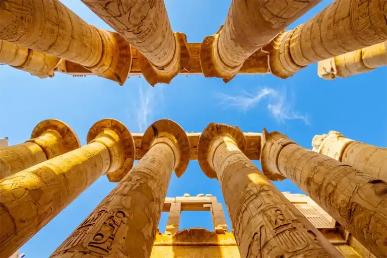 Imposing view looking up at the hieroglyph-carved stone columns of the Great Hypostyle Hall in Karnak Temple, Luxor. Clear blue sky visible between the massive columns.