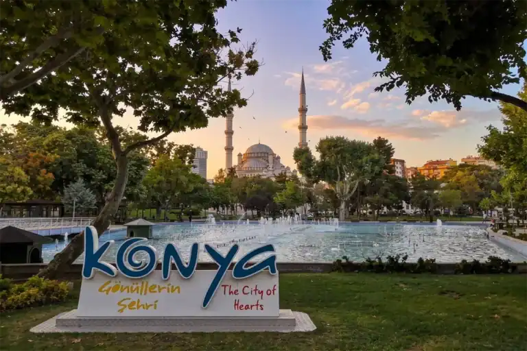 Konya park at sunset featuring a "Konya: The City of Hearts" sign, a water fountain, and the historic Sultan Selim Mosque in the background under a soft, cloudy sky.