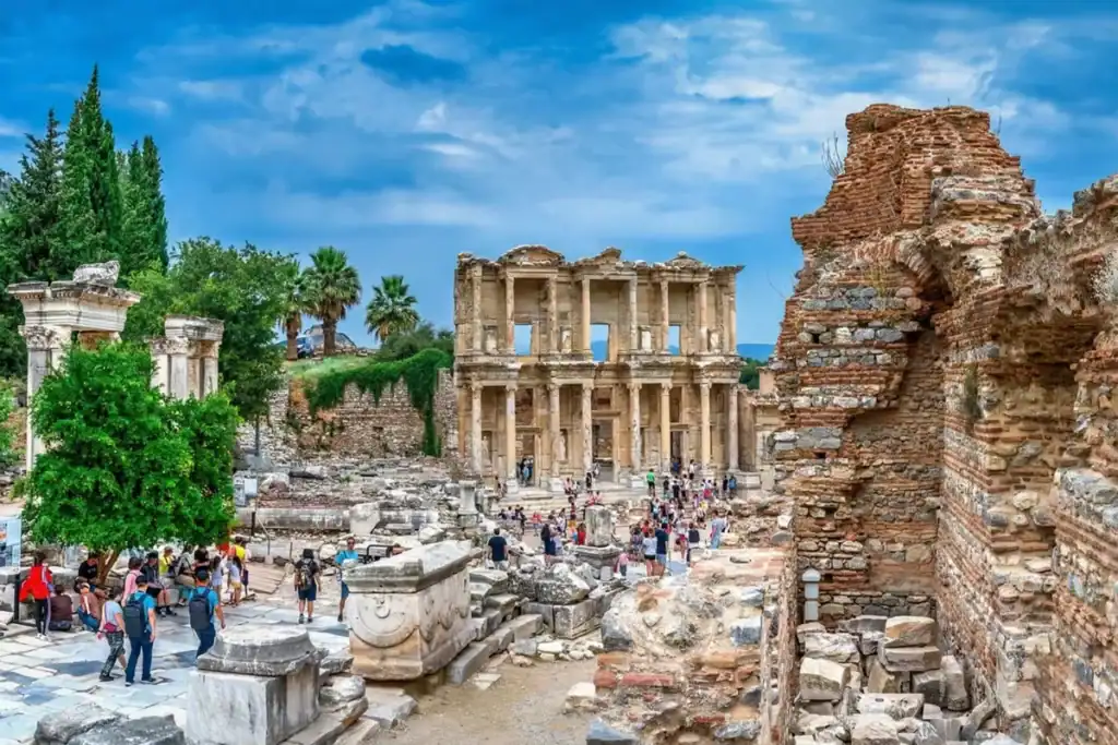 Visitors explore the historic Library of Celsus in the ancient city of Ephesus, Turkey, walking along marble pathways lined with columns and ruined stone structures under a cloudy sky.