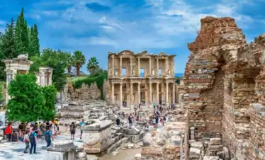 Visitors explore the historic Library of Celsus in the ancient city of Ephesus, Turkey, walking along marble pathways lined with columns and ruined stone structures under a cloudy sky.