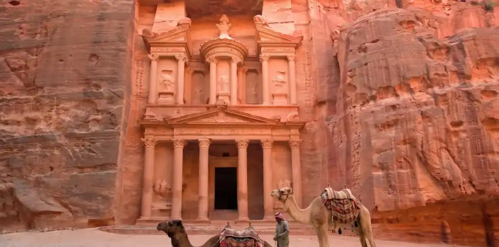A view of the ancient rock-cut Treasury (Al-Khazneh) facade in Petra, Jordan. In the foreground, a Bedouin man stands next to two camels—one sitting and one standing—all on sandy ground before the massive sandstone cliffs under warm light.