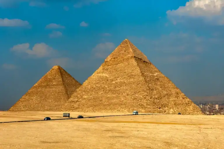 The majestic Pyramids of Giza standing in the Egyptian desert under a bright blue sky, with a road and vehicles in the foreground.