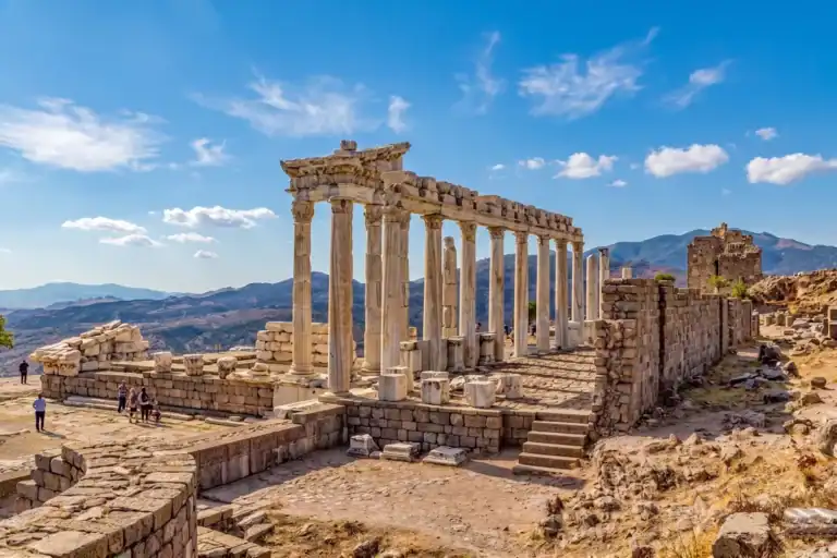A panoramic view of the ancient Temple of Trajan ruins at Pergamon, Turkey, featuring standing Corinthian columns on a hilltop overlooking a vast scenic valley and other structures.