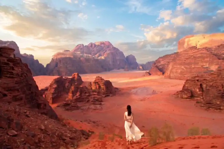 Back view of a woman in a white dress admiring the vast red sand and majestic rocky mountains of Wadi Rum, Jordan.