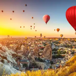 Hot air balloons drift over Cappadocia’s rock valleys and Göreme at sunrise, led by a vivid red balloon.