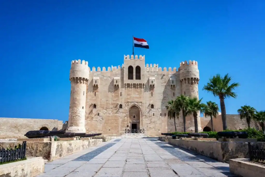 Citadel of Qaitbay in Alexandria, Egypt, with historic stone towers, palm trees, and the Egyptian flag under blue skies.