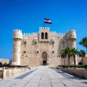 Citadel of Qaitbay in Alexandria, Egypt, with historic stone towers, palm trees, and the Egyptian flag under blue skies.