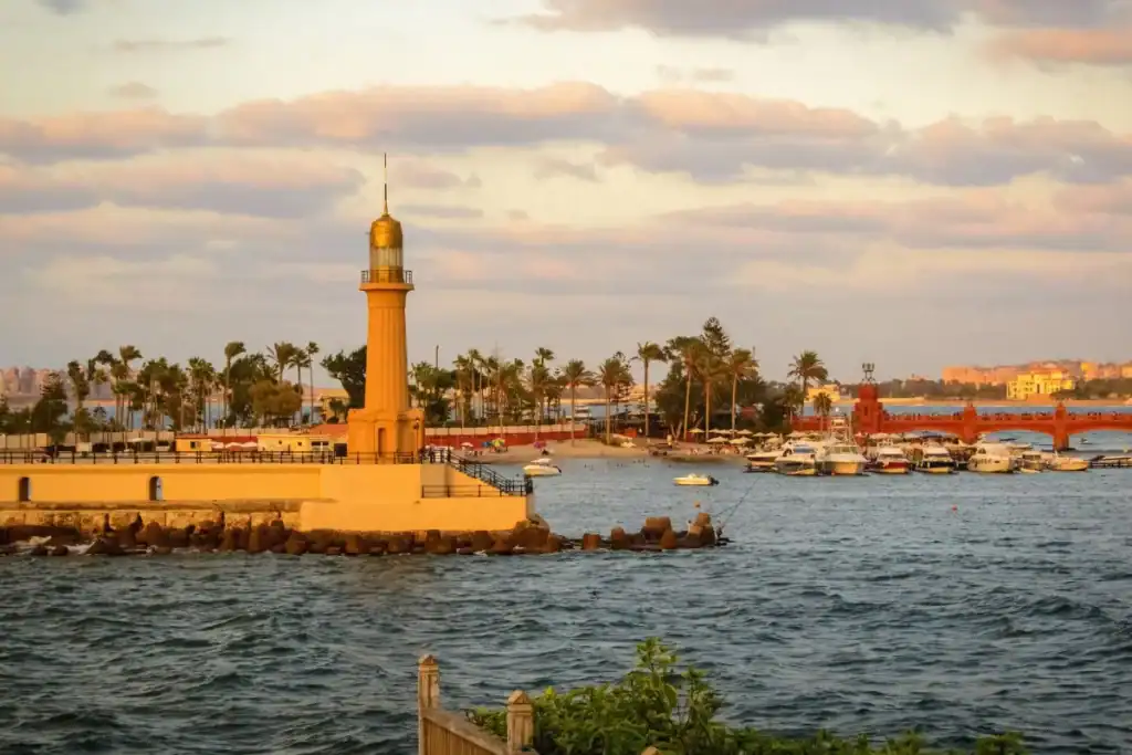 Lighthouse of Alexandria, Egypt, overlooking the Mediterranean with boats, palm trees, and a glowing sunset sky.