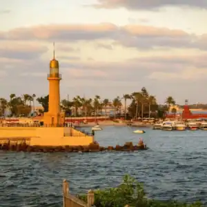 Lighthouse of Alexandria, Egypt, overlooking the Mediterranean with boats, palm trees, and a glowing sunset sky.