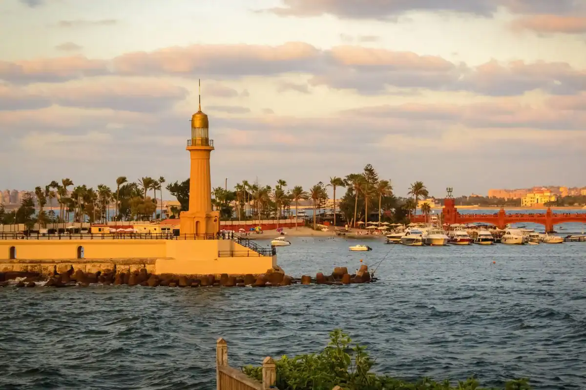 Lighthouse of Alexandria, Egypt, overlooking the Mediterranean with boats, palm trees, and a glowing sunset sky.