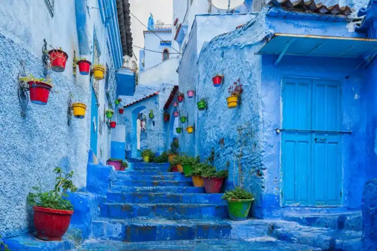 A narrow blue-painted street with stairs, colorful flower pots, and traditional doors in Chefchaouen, Morocco.