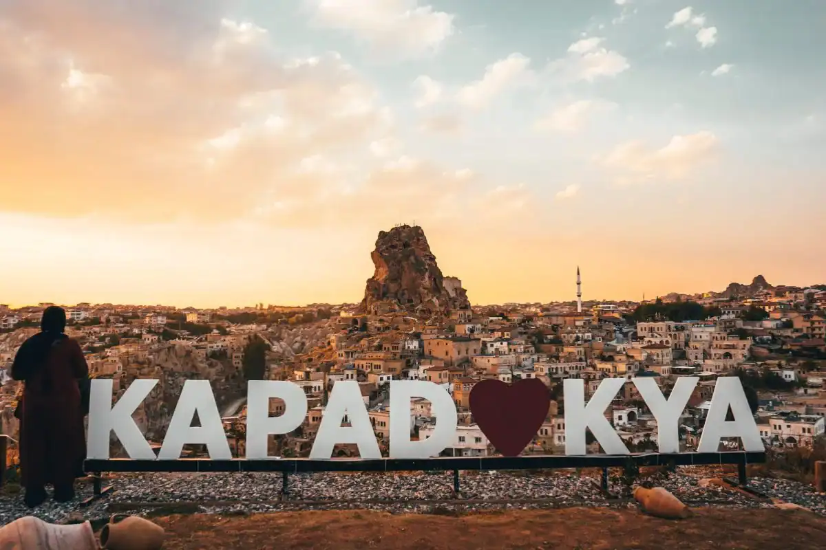 Cappadocia sunset view with Ortahisar rock castle, hillside homes, mosque minaret, and Kapadokya heart sign.