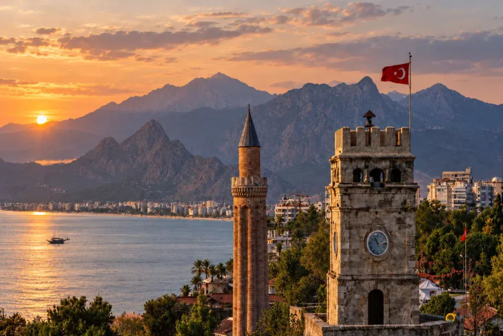 Antalya Clock Tower and historic minaret beside a Turkish flag, overlooking the Mediterranean Sea at sunset.